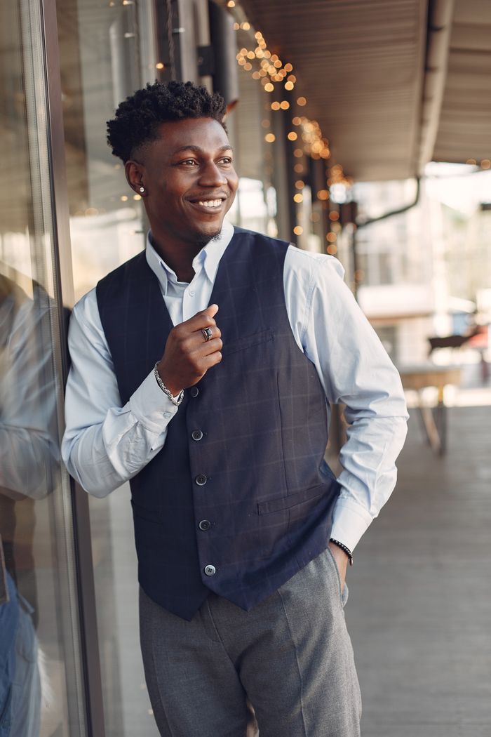 Services Smiling young handsome African American male standing on urban city street leaning on glass wall of cafeteria with hand in pocket and looking away