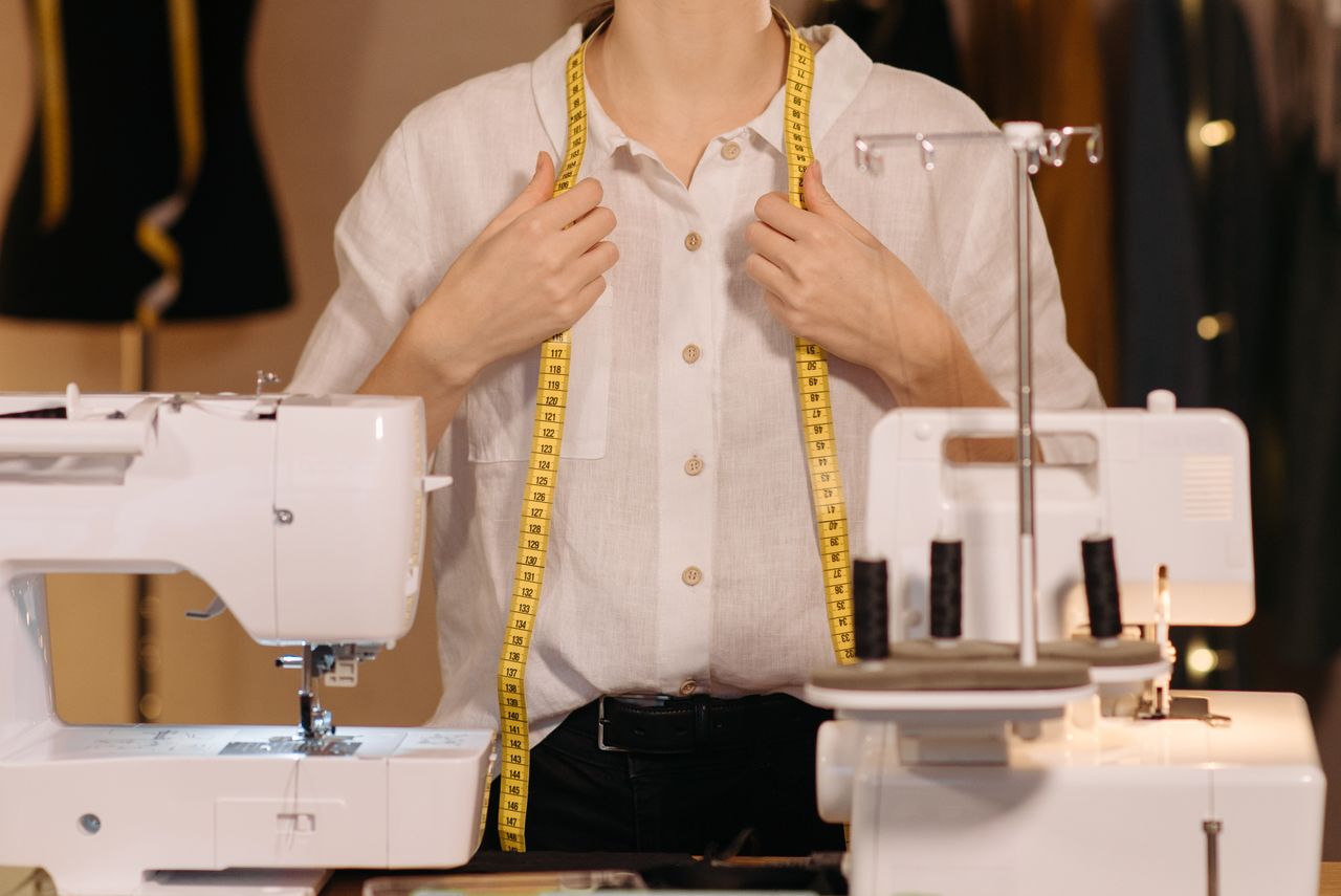 Services Tailor in a white shirt uses a measuring tape with sewing machines in a workspace.